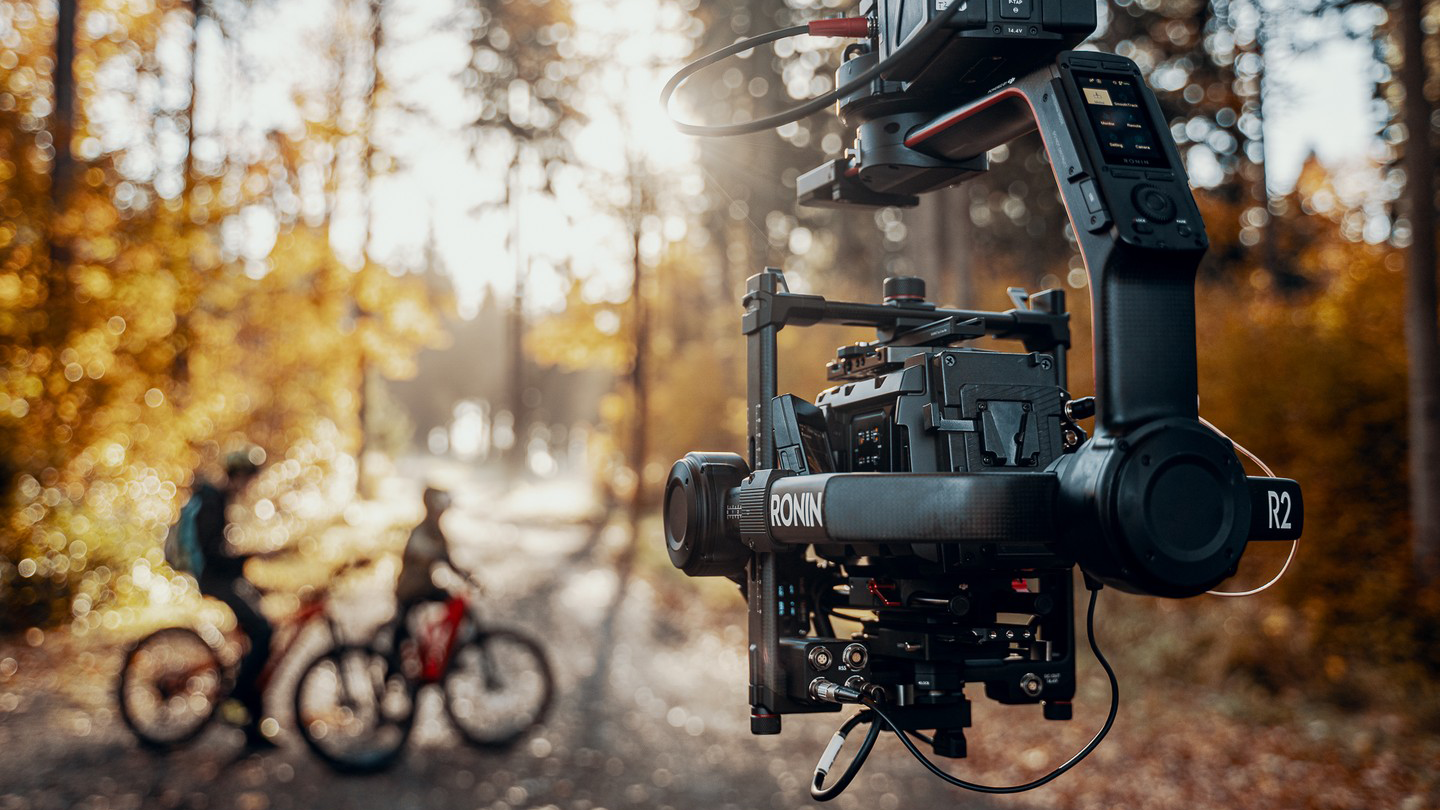 Stabilizator DJI Ronin 2 z kamerą na planie zdjęciowym w lesie, ujęcie outdoor z rowerzystami w tle